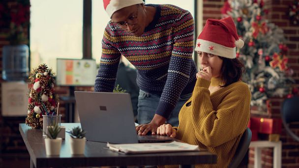 クリスマス休暇中のノートパソコンの情報を分析する多様な人々 xmasツリーとオフィスでのお祭り。季節の装飾や装飾が施されたデスクでコンピュータで作業する. - 写真・画像