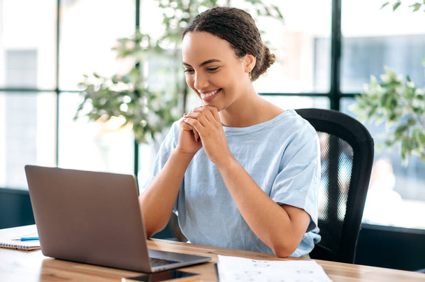 Udane pewność siebie piękna brazylijska lub latynoska kobieta biznesu, pracownik biura, menedżer kreatywny, siedzi w miejscu pracy w nowoczesnym biurze, patrząc na laptopa, czytając dobre wieści, uśmiechając się szczęśliwie - Zdjęcie, obraz