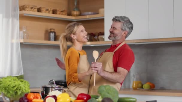 Funny spouses culinary. Happy laughing middle aged married man and woman dancing together and singing into big wooden spoon while cooking dinner at kitchen, slow motion - Footage, Video