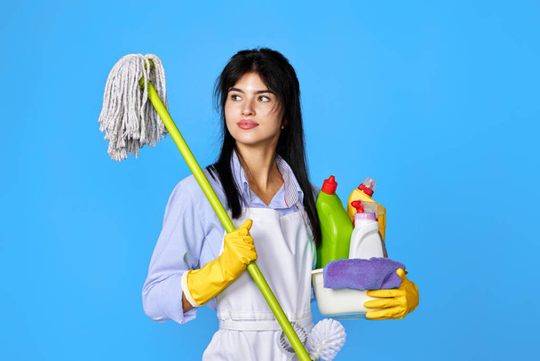 pretty woman in yellow rubber gloves and cleaner apron holding bucket of detergents and mop on blue background. - Foto, immagini