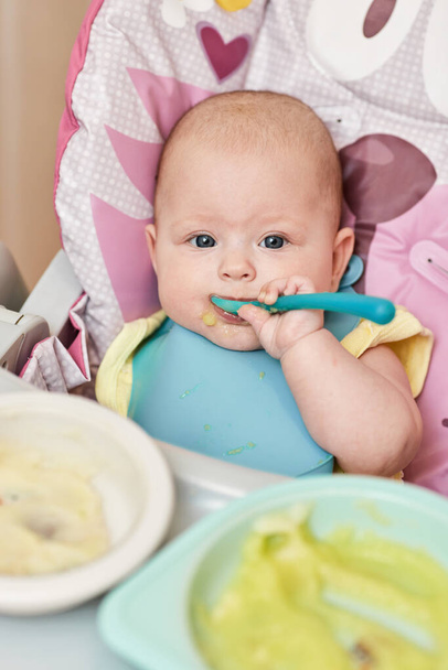 cute baby girl eating with spoon on baby chair in kitchen - Zdjęcie, obraz
