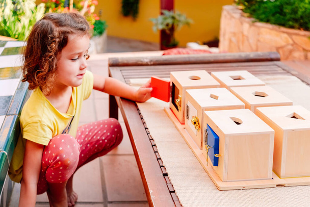 Ένα κορίτσι μαθαίνει χρησιμοποιώντας τα χέρια της να παίζει με ξύλινα κουτιά με κλειδαριές με την παιδαγωγική Montessori. - Φωτογραφία, εικόνα