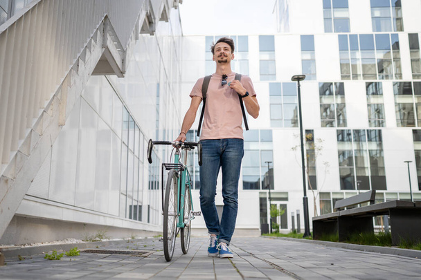 een man jong volwassen blanke man wandelen door het moderne gebouw met zijn fiets in de dag volledige lengte vooraanzicht kopieerruimte mannelijke student in de voorkant van de universiteit casual lifestyle concept - Foto, afbeelding