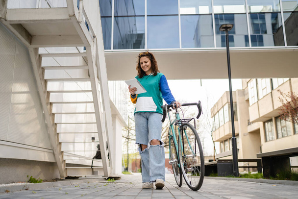 Bir yetişkin kadın, modern binanın önünden bisikletle geçen bir beyaz kadın. Üniversite önündeki uzay fotokopi uzmanı kadın öğrenci. Günlük yaşam tarzı kavramı dijital tablet taşıyor. - Fotoğraf, Görsel