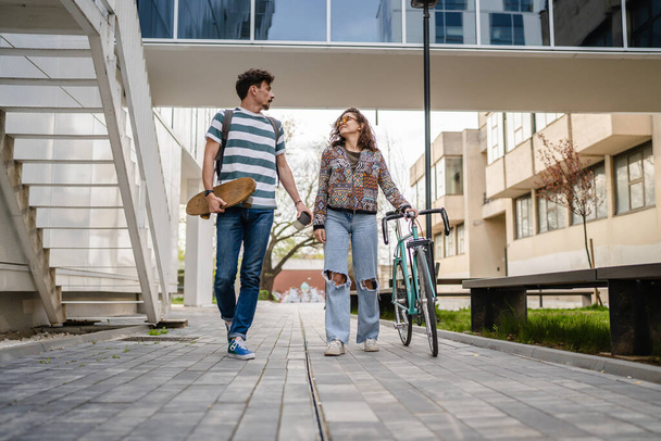 Two people young adult couple man and woman boyfriend and girlfriend or students friends walk by the building or university in day with bicycle and skateboard modern lifestyle concept copy pace - Zdjęcie, obraz