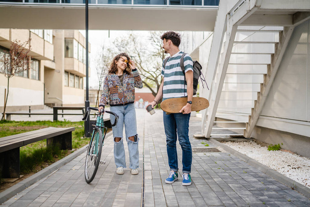 2人の若い大人のカップルの男性と女性のボーイフレンドとガールフレンドや学生の友人が自転車やスケートボード現代的なライフスタイルのコンセプトコピーペースで建物や大学で一日を歩く - 写真・画像