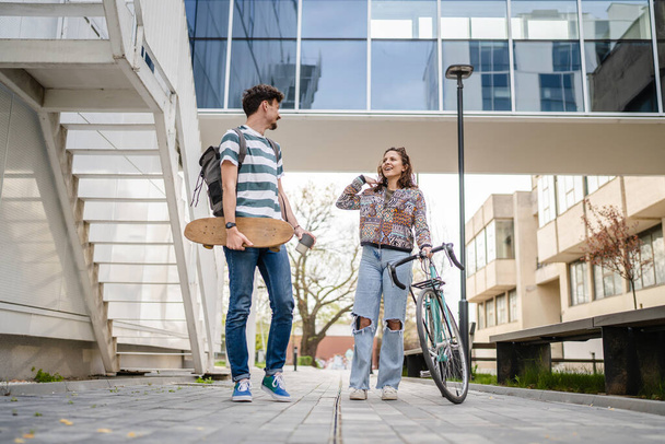 Two people young adult couple man and woman boyfriend and girlfriend or students friends walk by the building or university in day with bicycle and skateboard modern lifestyle concept copy pace - Photo, Image