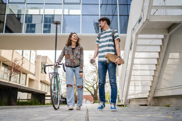 Two people young adult couple man and woman boyfriend and girlfriend or students friends walk by the building or university in day with bicycle and skateboard modern lifestyle concept copy pace - Фото, изображение