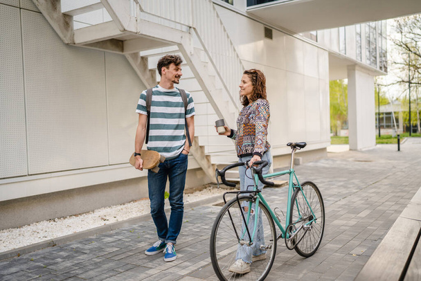 Two people young adult couple man and woman boyfriend and girlfriend or students friends walk by the building or university in day with bicycle and skateboard modern lifestyle concept copy pace - Foto, imagen