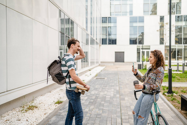 nuori aikuinen mies seisoo edessä modernin rakennuksen, kun hänen tyttöystävänsä nainen ottaa valokuvan matkapuhelimen valokuvaus opiskelija päivä ystävyys tai pari liimaus ja dating käsite - Valokuva, kuva