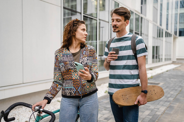 Δύο άτομα νέοι ενήλικες ζευγάρι άντρας και γυναίκα φίλος και φίλη ή οι φίλοι φοιτητές μιλούν από το κτίριο ή το πανεπιστήμιο την ημέρα με ποδήλατο και skateboard σύγχρονο τρόπο ζωής έννοια ρυθμό αντιγραφή - Φωτογραφία, εικόνα