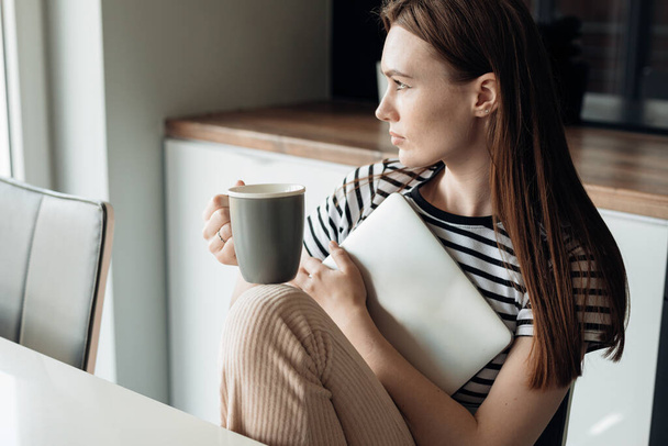 Młoda brązowłosa kobieta pijąca kawę i trzymająca biały laptop w ręku w kuchni, wyglądająca przez okno. Przygotowuję się do spotkania. E-learning, nowe zajecie, zdalna praca i edukacja. Bezsenność - Zdjęcie, obraz