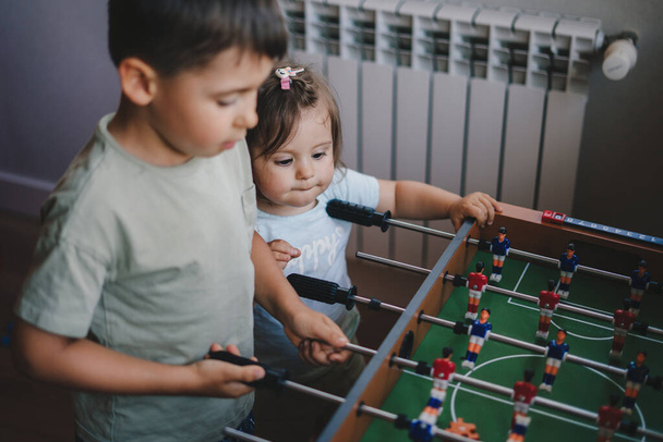 Χαρούμενα μικρά παιδιά που παίζουν επιτραπέζιο ποδόσφαιρο στο σπίτι, διασκεδάζουν με το επιτραπέζιο ποδόσφαιρο, σε εσωτερικούς χώρους. Χρόνος απομόνωσης του ιού Κορόνα. Ευτυχισμένη οικογένεια. Διασκεδαστική οικογένεια - Φωτογραφία, εικόνα