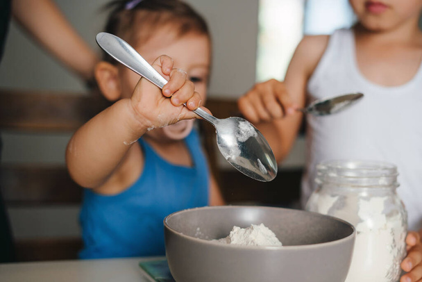 Linda niña añadiendo con una cuchara ingredientes en un tazón mientras se hace la masa para cocinar panadería en la cocina. Preparando pasteles caseros, expresando emociones positivas - Foto, imagen