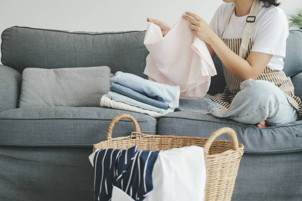 Young woman folding clothes while sitting on sofa in living room at home. Woman doing housechores, laundry, and folding clothes at home. Housekeeping concept. - Fotoğraf, Görsel