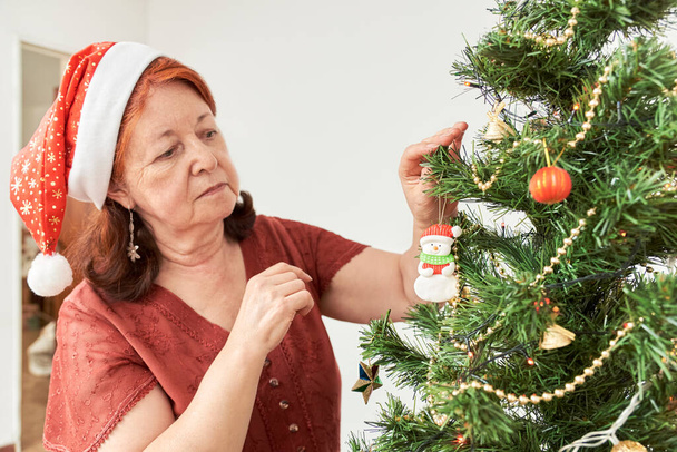 Femme âgée hispanique décorant un sapin de Noël à la maison portant un bonnet rouge du Père Noël, accrochant un ornement de bonhomme de neige sur une branche. - Photo, image