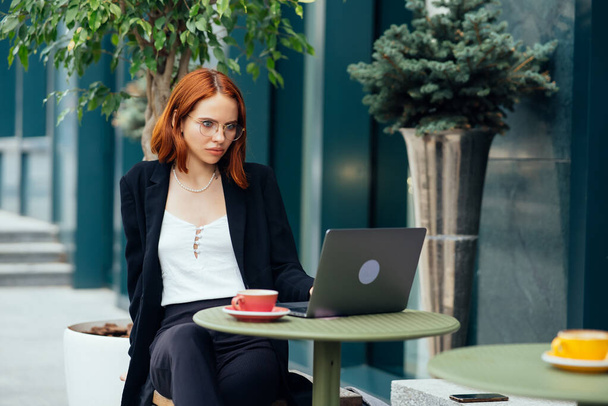 Beautiful redhead woman working on laptop at outdoors cafe - Photo, Image