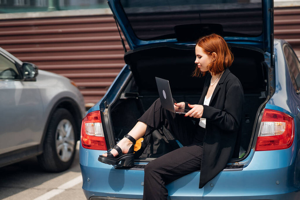 Mujer joven atractiva de negocios que trabaja en el ordenador portátil mientras está sentado en el maletero del coche mientras viaja - Foto, imagen