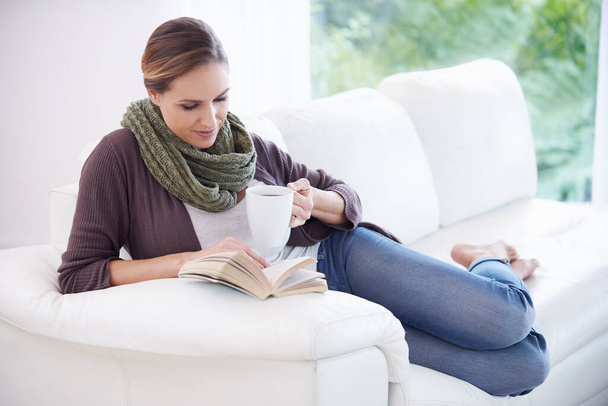 Bir sayfada kaybolan genç bir kadın elinde kitap ve bir fincan kahveyle kanepede dinleniyor. - Fotoğraf, Görsel