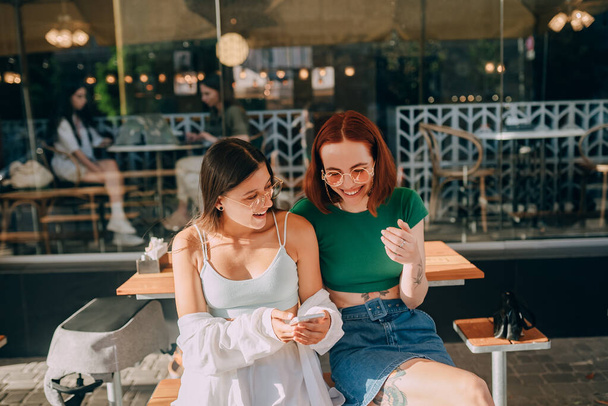 Kaksi innoissaan nuoret tytöt käyttävät matkapuhelimia istuessaan kahvilassa ulkona - Valokuva, kuva