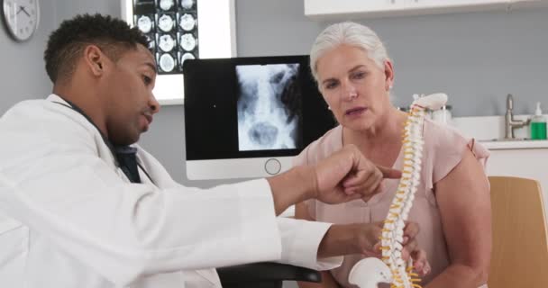 Portret młodego afrykańsko-amerykańskiego lekarza pokazujący pacjentowi model kręgosłupa kręgosłupa. zbliżenie w górę w senior kobieta odwiedzając kręgarz - Materiał filmowy, wideo
