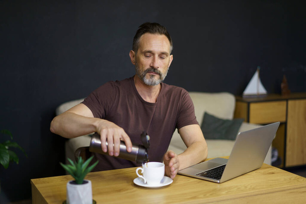 Bearded middle-age man pouring coffee from thermos to cup sitting at home working place with laptop - Zdjęcie, obraz