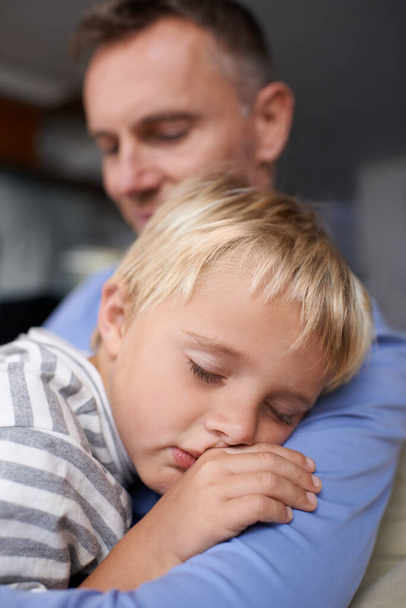 Slapen op papa 's schouder. Een volwassen vader die thuiskomt van zijn werk en zijn uitgeputte kleine jongen knuffelt - Foto, afbeelding