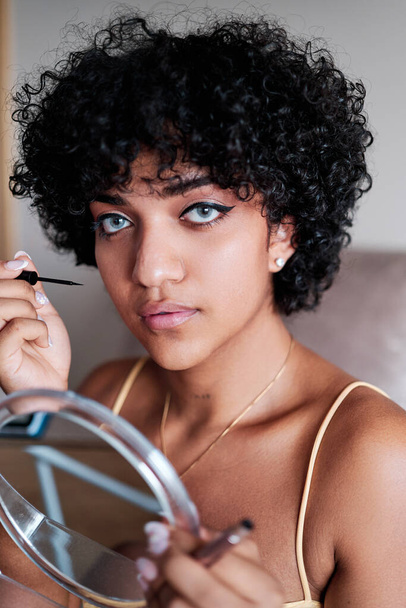 Transgender-Frau mit lockigem Haar blickt in die Kamera, während sie einen Spiegel hält und sich zu Hause schminkt. - Foto, Bild
