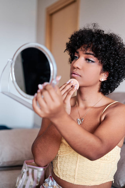 Transgender woman with curly hair holding and looking at a small mirror while applying compact powder makeup on her face at home. - Fotó, kép