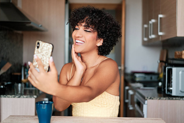 Joyful transgender woman smiling while taking selfies with a mobile phone sitting in the kitchen at home. Technology concept. - Fotó, kép