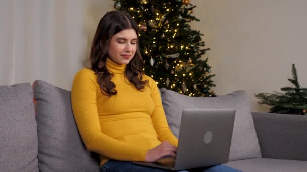笑顔の若い女性は、バックグラウンドクリスマスツリー上の親友とノートパソコンのオンラインチャットを使用しています。正の美しい女性の距離作品のためにコンピュータタイピングテキスト上のキーボード座って上のソファに家 - 映像、動画