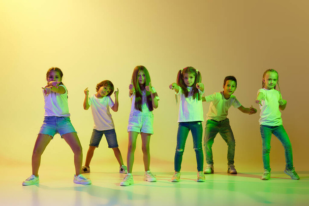 Street style. Stylish children, little girls and boys in casual style clothes dancing hip-hop isolated on green background in yellow neon light. Concept of music, fashion, art, modern contemp dance - Foto, afbeelding