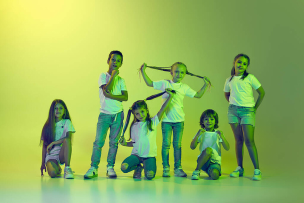 Hip-hop dancing. Group of children, little girls and boys in sportive casual style clothes dancing in choreography class isolated on green background in yellow neon. Concept of music, fashion, art - Zdjęcie, obraz