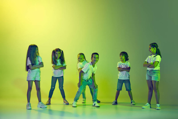Hip-hop dancing. Group of children, little girls and boys in sportive casual style clothes dancing in choreography class isolated on green background in yellow neon. Concept of music, fashion, art - Foto, Imagen