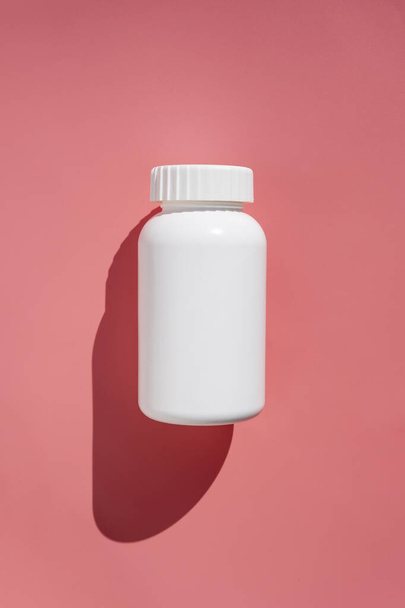 白いプラスチック製の薬瓶、ビタミンや食品サプリメット。太陽の光と影のピンクの背景に薬の丸薬でコンテナ - 写真・画像