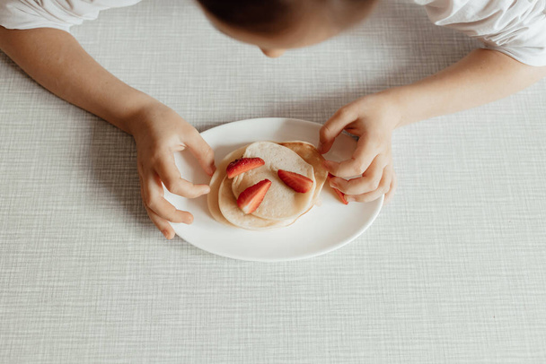Aç küçük kız çilekli panckaces yiyor. Aile kahvaltısı kavramı. Üzerinde çilekli krep olan bir tabakla çocuk elleri. - Fotoğraf, Görsel