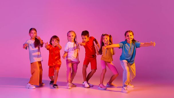 Modern choreography. Dance group of happy, active little girls and boys in bright clothes in action isolated on pink background in neon. Concept of emotions, music, fashion, art, childhood, hobby. - Foto, Bild