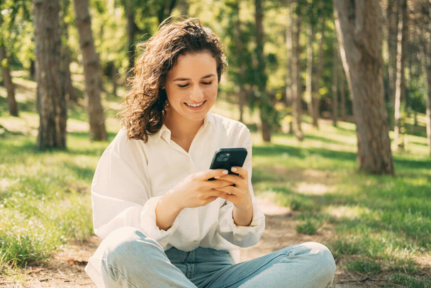 Foto da jovem mulher feliz sorrindo e digitando no smartphone ao ar livre no parque durante o dia ensolarado. - Foto, Imagem