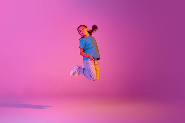 Freestyle. Beautiful active little girl, kid jumping high, dancing isolated over pink background in neon. Kids emotions, news, challenges, childhood, education concept. Copy space for ad - Foto, Imagem