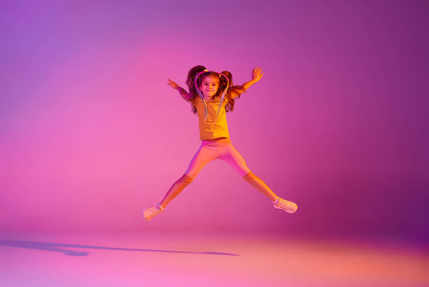 Flight. Expressive little girl, kid in casual bright clothes jumping isolated over pink background in neon. Kids emotions, news, challenges, childhood, education concept. Copy space for ad - Photo, image