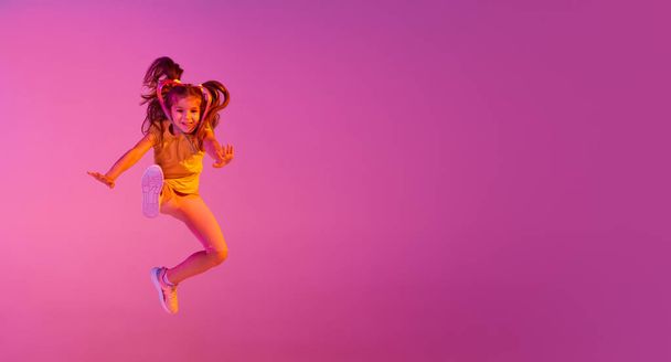 Freestyle. Beautiful active little girl, kid jumping high, dancing isolated over pink background in neon. Kids emotions, news, challenges, childhood, education concept. Copy space for ad - Fotografie, Obrázek