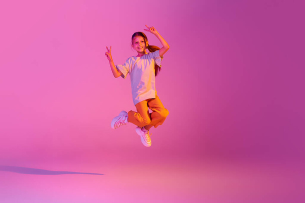Freestyle. Beautiful active little girl, kid jumping high, dancing isolated over pink background in neon. Kids emotions, news, challenges, childhood, education concept. Copy space for ad - Valokuva, kuva