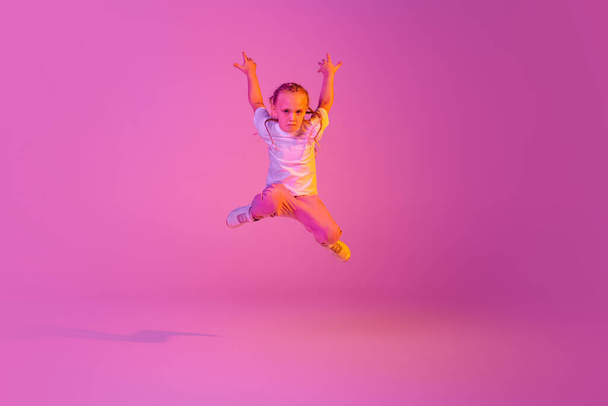 Flight. Expressive little girl, kid in casual bright clothes jumping isolated over pink background in neon. Kids emotions, news, challenges, childhood, education concept. Copy space for ad - Фото, изображение