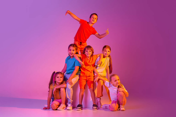 Modern choreography. Dance group of happy, active little girls and boys in bright clothes in action isolated on pink background in neon. Concept of emotions, music, fashion, art, childhood, hobby. - Фото, изображение
