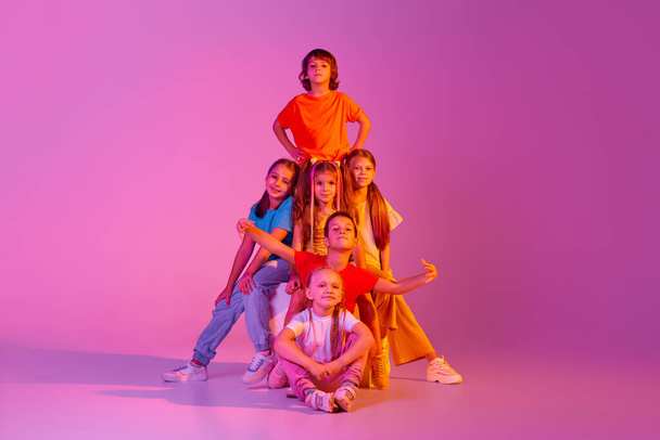 Friends, classmates. Happy, joyful kids, school age girls and boys in bright clothes posing isolated on pink background in neon. Concept of emotions, music, fashion, art, childhood, hobby. - Фото, изображение