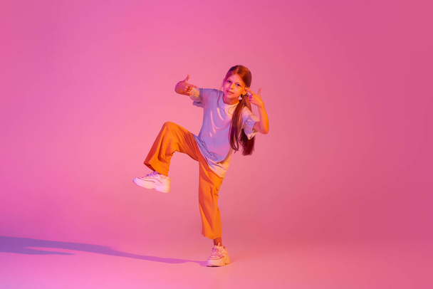 Hip-hop dance. Portrait of little girl, kid in casual bright clothes dancing isolated over pink background in neon. Kids emotions, news, challenges, childhood, education concept. Copy space for ad - Foto, imagen