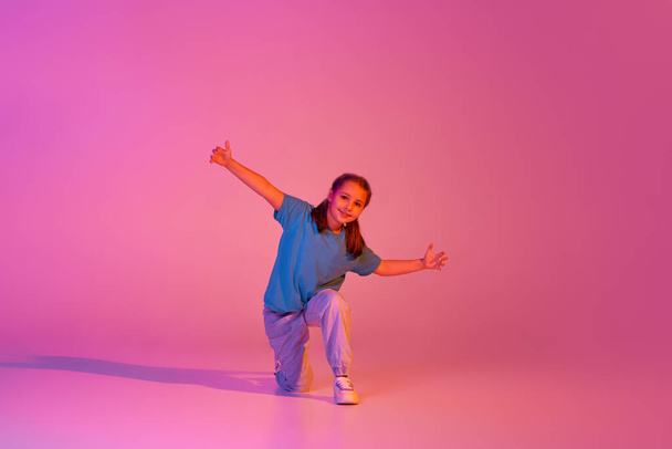 Hip-hop dance. Portrait of little girl, kid in casual bright clothes dancing isolated over pink background in neon. Kids emotions, news, challenges, childhood, education concept. Copy space for ad - Foto, immagini