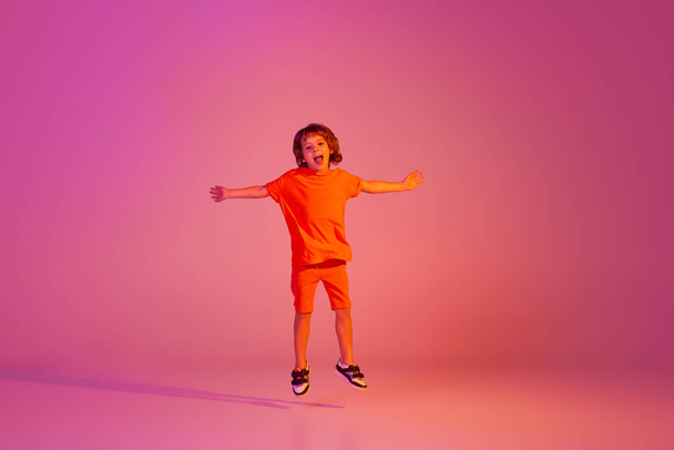 Portrait of caucasian little boy, kid in casual bright clothes dancing isolated over pink background in neon. Kids emotions, news, challenges, childhood, education concept. Copy space for ad - Fotoğraf, Görsel