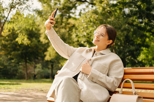 Photo of young smiling woman taking selfie outdoor in park during autumn. - Фото, зображення
