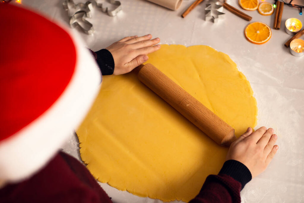 Niño en sombrero de navidad saca la masa con rodillo para formar galletas. Ambiente navideño, familia horneando juntos. - Foto, imagen